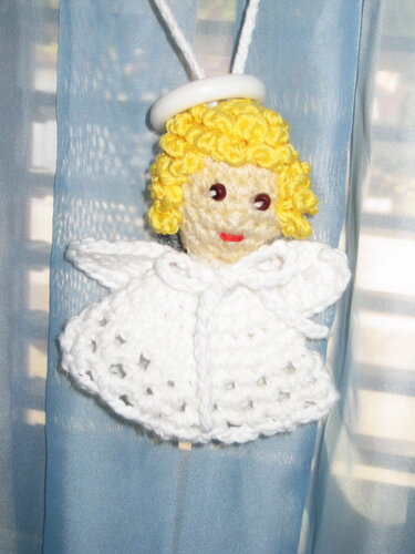 Crocheted amigurumi angel ornament with yellow curly hair, white gown, and halo hanging against a blue fabric background.