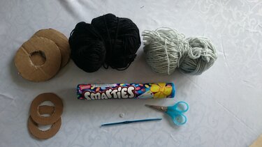 Bastelmaterialien mit schwarzem und hellgrauem Garn, Pappringen, Häkelnadel, Schere und Knopf auf hellem Untergrund.