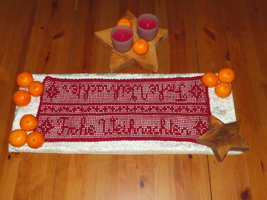 Roter gehäkelter Tischläufer mit den Schriftzügen „Frohe Weihnachten“ und „Schöne Feiertage“, dekoriert mit Mandarinen und Kerzen auf Holzsternen auf einem Holztisch.