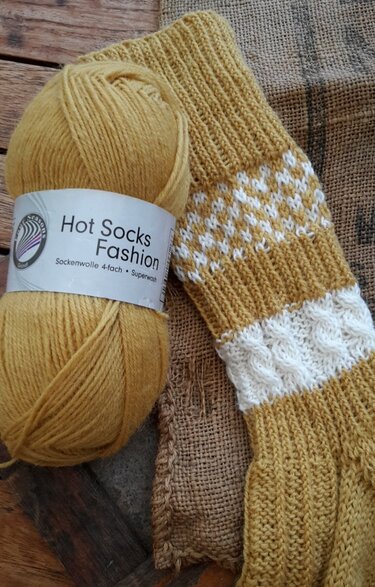 Detailaufnahme von senfgelbem Sockengarn und einer Socke mit Rippenbündchen sowie weißem Jacquard- und Zopfmuster