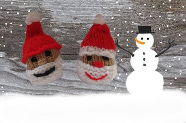 Zwei gestrickte Weihnachtsmannköpfe mit roten Mützen und weißen Bärten vor Holzuntergrund neben gezeichnetem Schneemann