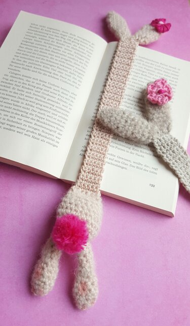 Pastellfarbenes gehäkeltes Häschen-Lesezeichen mit Bommel und Häkelblüten auf offenem Buch vor rosafarbenem Hintergrund