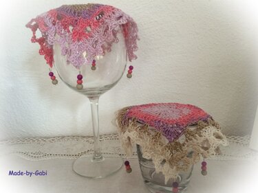 Zwei gehäkelte Glasabdeckungen mit Perlen, eine auf einem Weinglas, die andere auf einem kleinen Glas