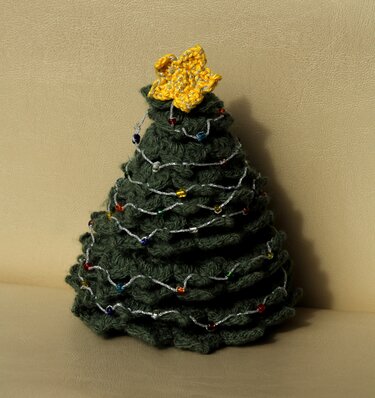 Dunkelgrüner handgehäkelter Mini-Weihnachtsbaum mit gelbem Stern und silberner Perlenkette vor beigem Hintergrund.
