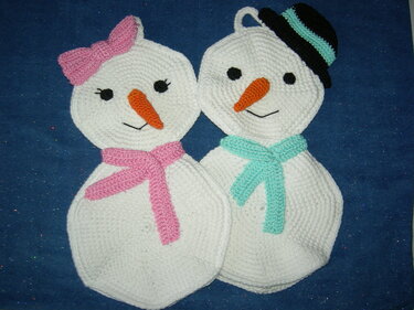 Zwei gehäkelte Schneemann-Topflappen auf blauem Hintergrund; links mit rosa Schleife und Schal, rechts mit schwarzem Hut und türkisfarbenem Schal.
