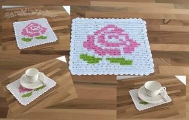Weißer, quadratischer gehäkelter Untersetzer mit rosa Rose und grünen Blättern auf Holzfläche