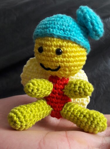 Kleine bunte Amigurumi-Figur mit gelbem Körper, blauer Mütze und grünen Gliedmaßen auf einer Hand