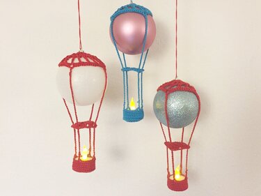 Drei gehäkelte Heißluftballon-Ornamente hängen an roten und blauen Fäden, mit leuchtenden LED-Teelichtern in den gehäkelten Körben.