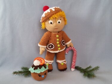 Gehäkelte Amigurumi-Puppe im Lebkuchen-Stil mit brauner Mütze und Jacke, daneben Zuckerstange und Cupcake