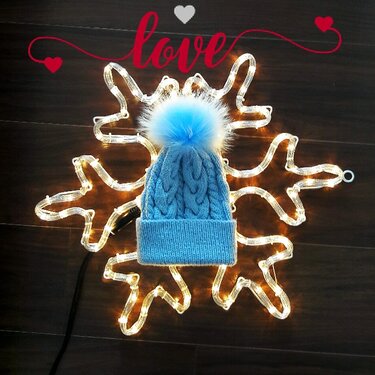 Hellblaue Strickmütze mit Zopfmuster und großem hellblauen Bommel auf einer beleuchteten Schneeflocken-Lichterkette; rote 'love'-Grafik oben.
