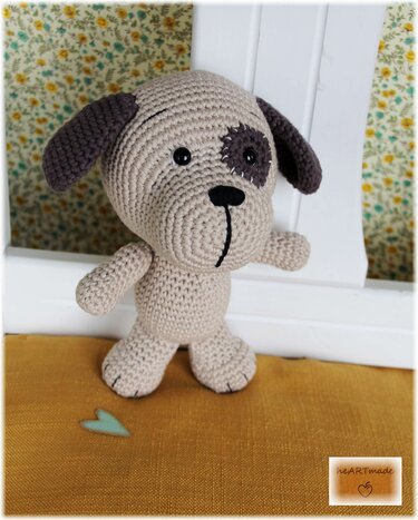 Beiger gehäkelter Amigurumi-Hund mit dunklen Ohren und Augenpatch auf senffarbenem Stoff
