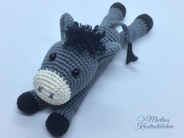 Handgehäkelter grauer Amigurumi-Esel mit hellbeiger Schnauze und schwarzen Hufen, liegend auf hellem Untergrund.