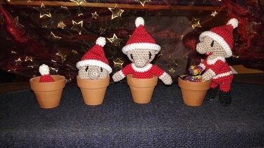 Vier gehäkelte Weihnachtswichtel mit roten Mützen, drei in kleinen Tontöpfen, vor rotem Stoff mit goldenen Sternen.