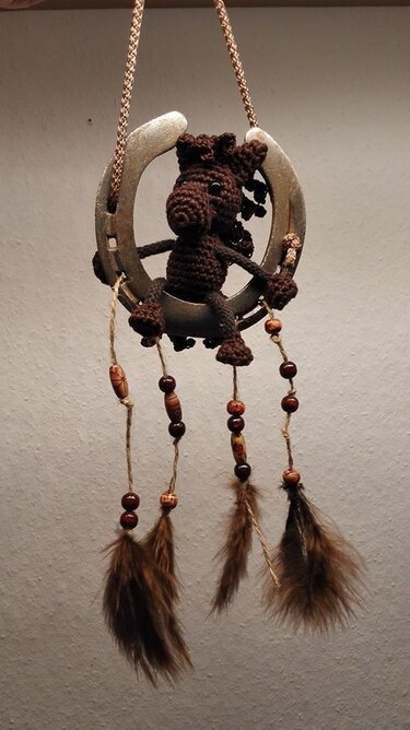 Braunes gehäkeltes Amigurumi-Pferd sitzt in einem Hufeisen, darunter hängen Perlen und braune Federn.