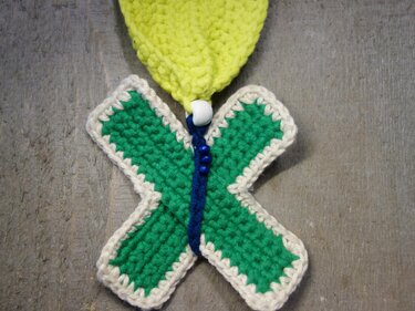 Gehäkelter grüner Schmetterling mit cremefarbener Umrandung, blauen Perlen und gelbem Aufhänger auf Holz.