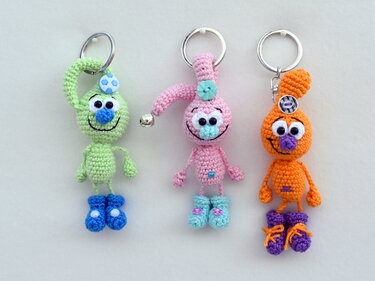 Drei gehäkelte Amigurumi-Schlüsselanhänger in Grün, Pink und Orange mit verschiedenen Details an Metallringen.