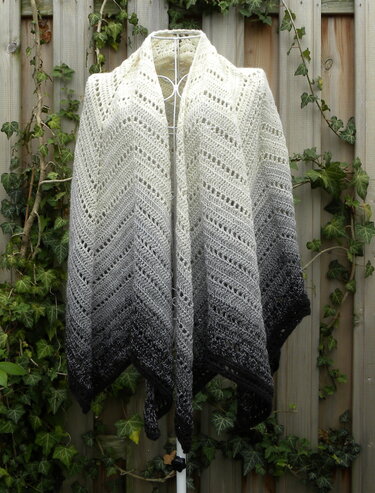 Gehäkelter Poncho in Creme, Grau und Schwarz mit Zickzack-Lochmuster an einer weißen Kleiderpuppe vor einem Holzzaun mit Efeu.