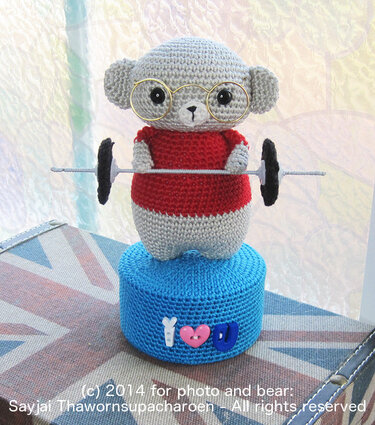 Kleiner Amigurumi‑Teddy mit Brille, rotem Oberteil und Hantel auf rundem blauem Sockel