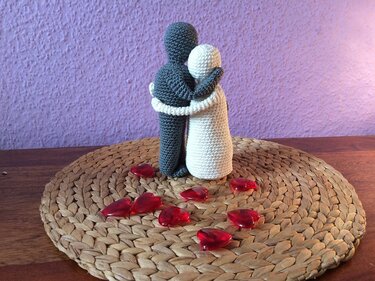 Gehäkelte Amigurumi-Figuren in Creme und Grau umarmen sich als Häkelidee, dazu rote Herzperlen