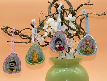 Vier gehäkelte Osteranhänger (Biene, Küken, Marienkäfer, Hase) an einem blühenden Zweig über einer grünen Vase