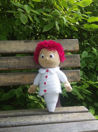 Gehäkelte Amigurumi-Puppe mit pinken Haaren und weißem Outfit auf einer Holzbank vor grünem Laub