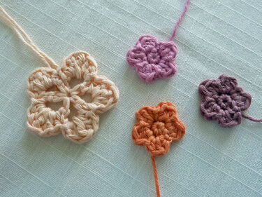 Vier gehäkelte Blumen-Applikationen in Creme, Flieder, Mauve und Orange auf hellem Stoff