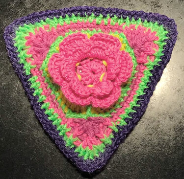 Dreieckige gehäkelte Applikation mit 3D-pinker Blüte, grünen und lila Reihen