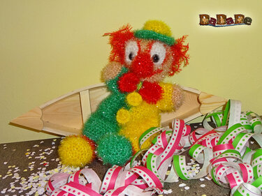 Gehäkelter bunter Clown mit rotem Haar, gelbem Hut und grüner Schärpe in einem Holzschiffchen, umgeben von Luftschlangen und Konfetti.