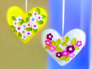 Zwei gehäkelte Herz-Anhänger, gelb und weiß, mit aufgenähten bunten Blumen und grünen Blättern.