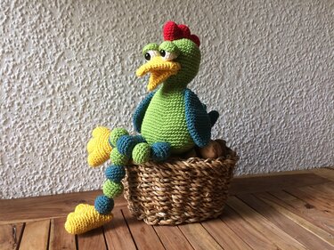 Gehäkelter grüner Amigurumi-Hahn mit rotem Kamm und gelbem Schnabel sitzt in einem geflochtenen Korb auf einem Holztisch.