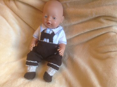 Babypuppe mit brauner gestrickter Latzhose, weißen Söckchen und weißem Kurzarmshirt auf beiger Decke