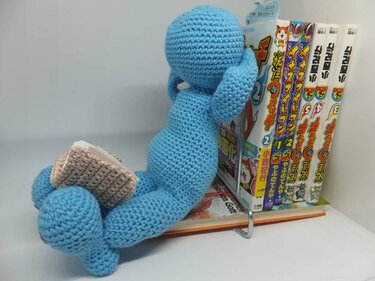 Blaue gehäkelte Amigurumi-Lesepuppe in liegender Pose hält ein kleines Buch und stützt Bücher im Regal
