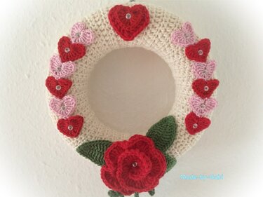 Cremefarbener gehäkelter Kranz mit roten und rosa Herzen und roter Häkelrose mit grünen Blättern