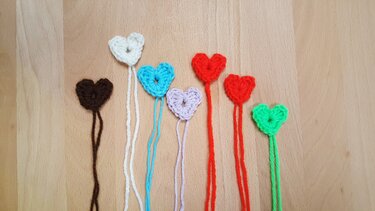 Sieben gehäkelte Herzen in Braun, Weiß, Hellblau, Lavendel, zwei Rottönen und Grün mit Garnenden auf hellem Holz