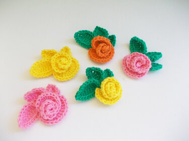 Fünf kleine gehäkelte Rosenapplikationen in Gelb, Orange und Pink mit grünen Blättern auf weißem Hintergrund