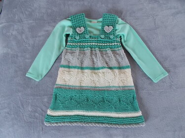 Gestricktes Mädchen-Latzkleid in Türkis, Grau und Creme mit Herzapplikationen auf den Trägern, liegend auf grauem Untergrund