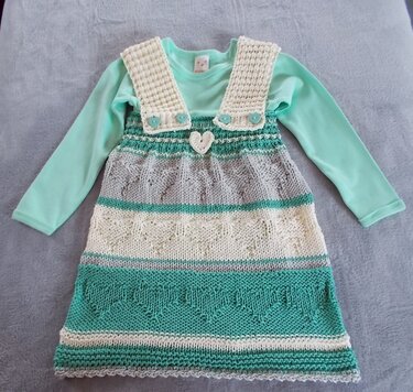 Gestricktes Kinderkleid in Mint, Grau und Creme mit Herzmustern und Knopfträgern, liegend auf hellem Untergrund