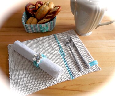 Gestricktes cremeweißes Tischset mit türkisen Akzenten, Serviettenring mit Blüte und gestreifter Brotkorb mit Backwaren.