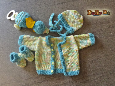 Von oben aufgenommenes handgestricktes Baby-Set in Pastellfarben mit türkisfarbener Kante, bestehend aus Jäckchen, Mütze, Schühchen und Rassel.