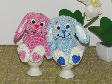 Zwei gehäkelte Hasen‑Eierwärmer (rosa und blau) in Eierbechern mit weißen Ohren und Herzpfoten