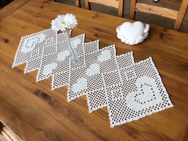 Weißer handgehäkelter Tischläufer mit Herzmotiven und gezacktem Rand auf Holztisch, dekoriert mit weißer Blume in Vase und herzförmiger Dekorskulptur.