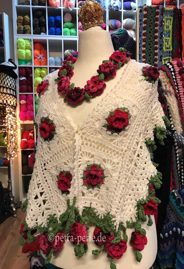 Weißer gehäkelter Poncho mit roten 3D‑Blüten auf Schaufensterpuppe vor Regal mit Wollgarnen