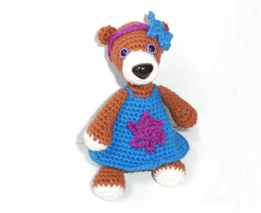 Gehäkelter brauner Teddybär mit weißer Schnauze, blauem Kleid und violettem Stirnband