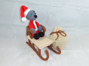 Gehäkelter grauer Teddy in rotem Weihnachtskostüm auf einem beige-braunen Schlitten neben einem gehäkelten Sack