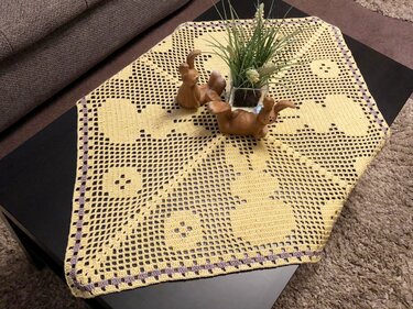 Gelber gehäkelter Tischläufer mit Hasen- und Eimotiven und lila Rand auf dunklem Tisch, dekoriert mit Keramikhasen und Pflanze