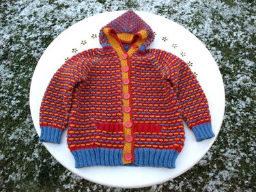 Mehrfarbige gestrickte Kinder-Kapuzenjacke mit Knopfleiste und Taschen, liegend auf weißem Tisch