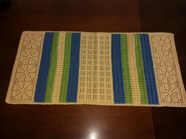 Gehäkelter rechteckiger Tischläufer in Beige mit blauen und grünen Streifen und Lochmuster auf dunklem Holztisch.