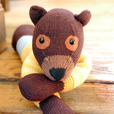 Close-up of a knitted amigurumi teddy bear with a brown head, tan snout and eye patches, black safety eyes, and a yellow striped body on a wooden floor