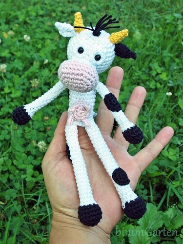 Weiße gehäkelte Amigurumi-Kuh mit schwarzen Flecken und langen Beinen, in einer Hand gehalten