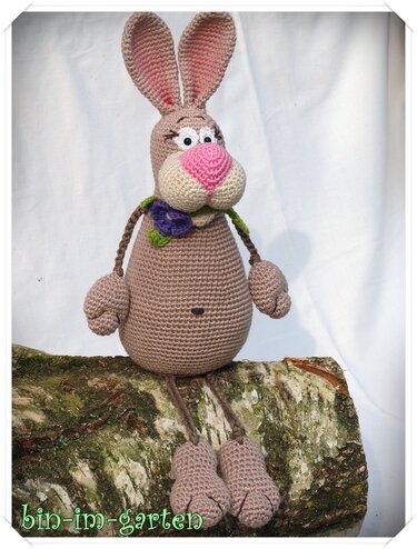 Amigurumi-Hase mit rundem Körper, geflochtenen Armen und langen Beinen, sitzend auf Stamm